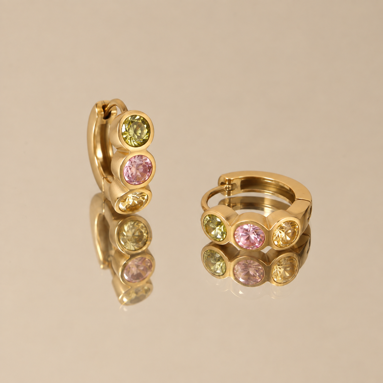 Pair of three stone gold huggie earrings with colorful zirconia on reflective surface