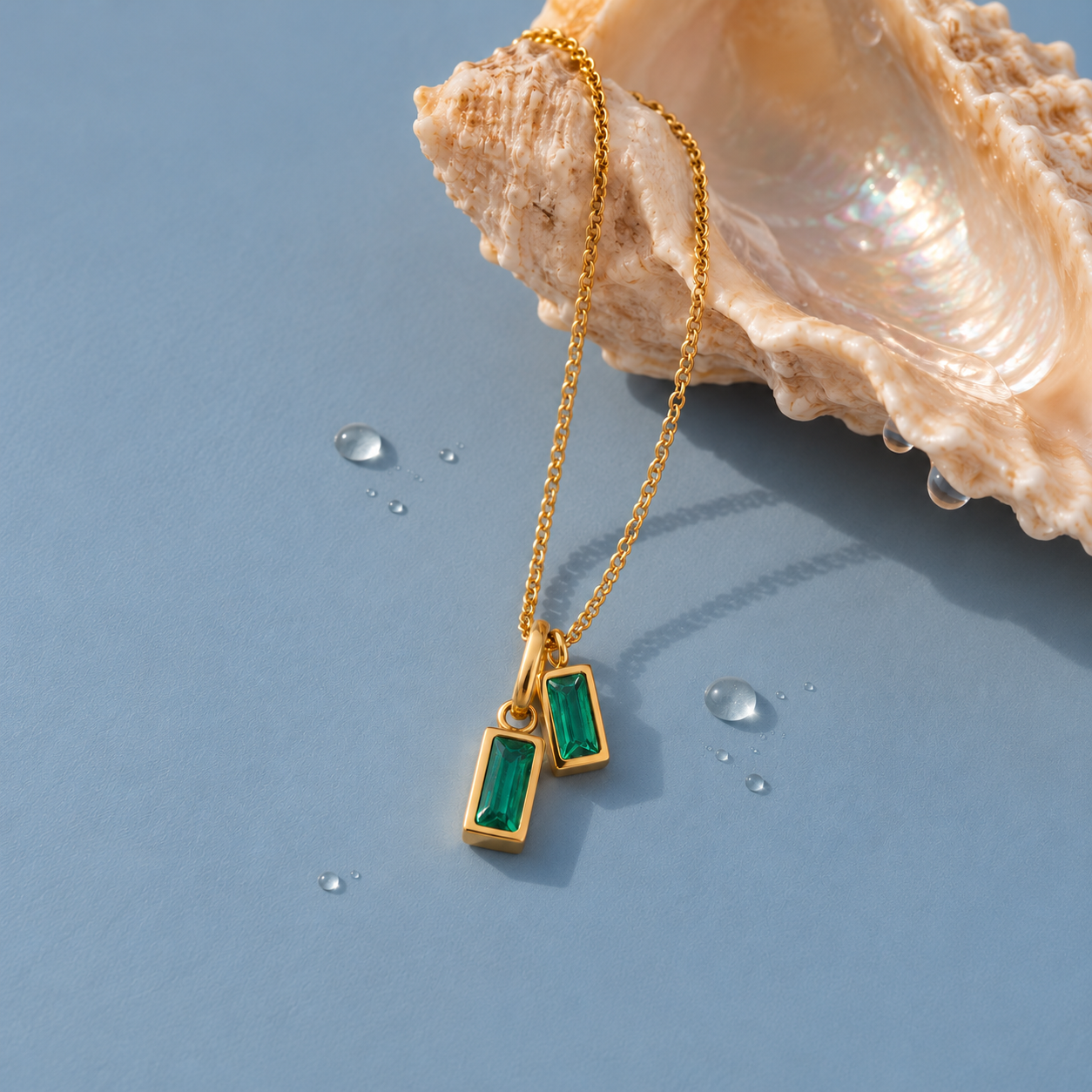Gold double emerald green pendant necklace draped over a pearlescent shell on a blue background with water droplets.
