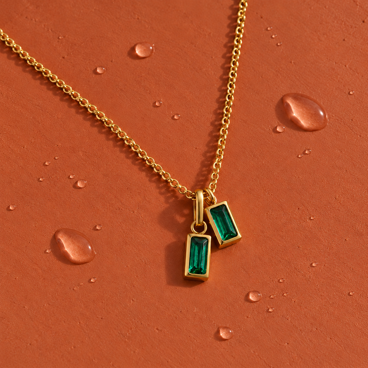 Gold necklace with two emerald green rectangular pendants on a terracotta background with water droplets.