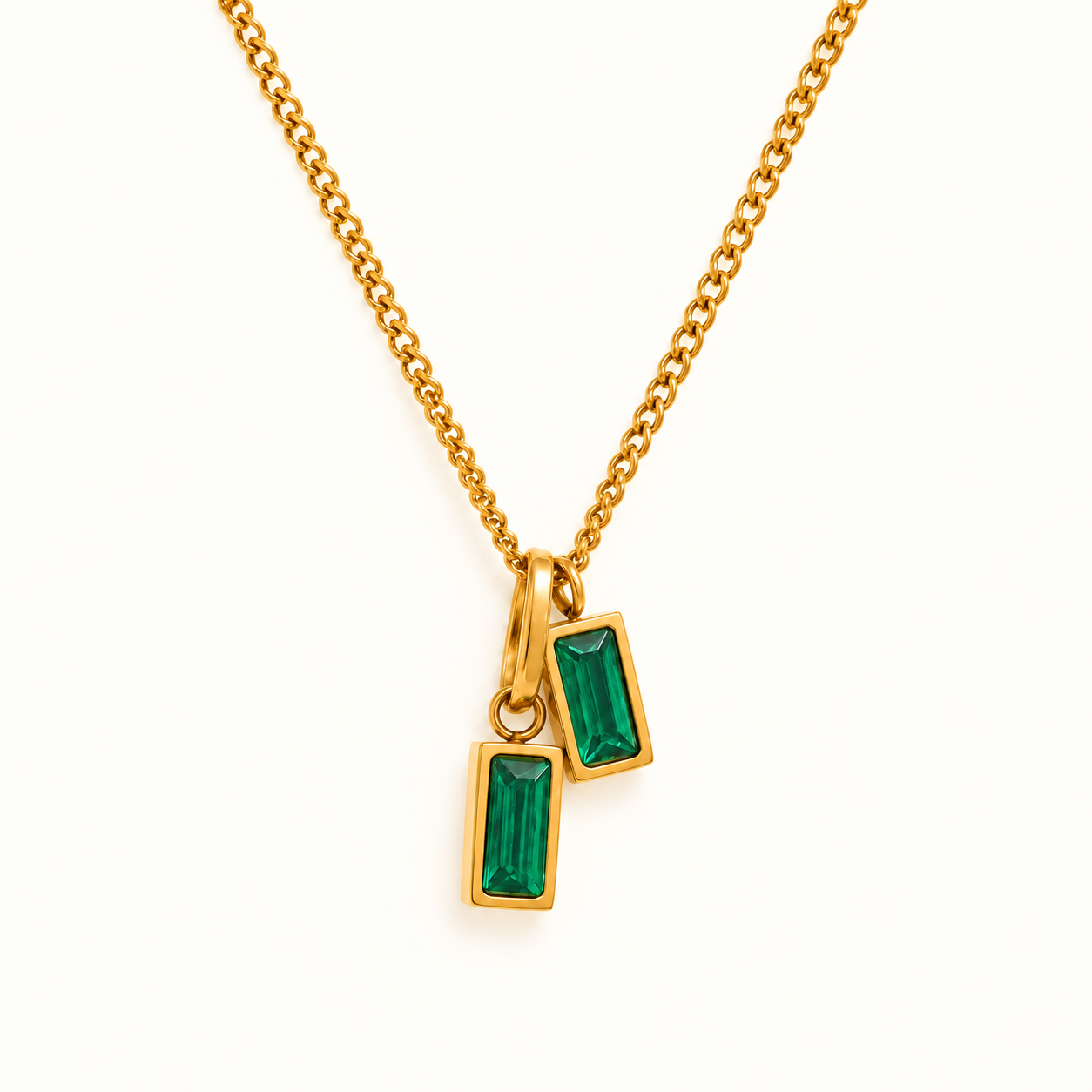 Gold necklace with two staggered emerald green baguette pendants on a plain ivory background.