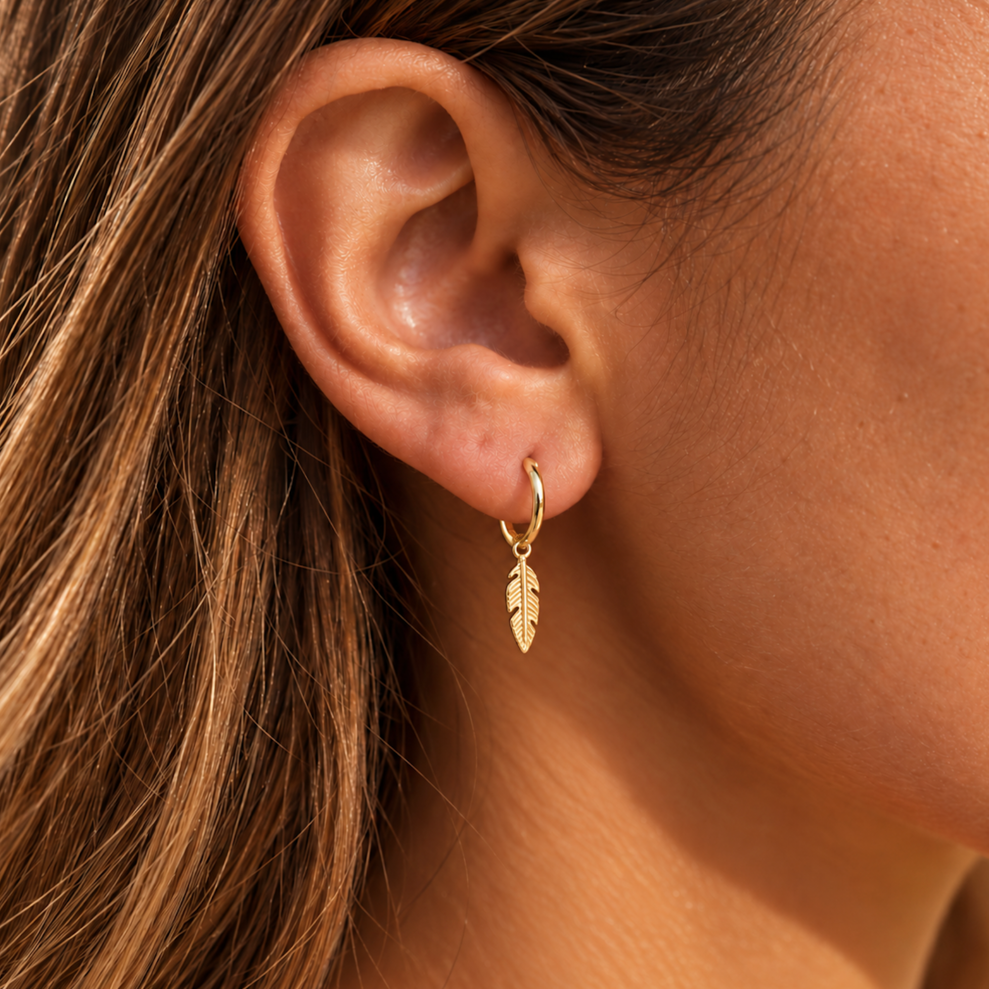Close-up of a gold feather charm huggie earring worn on the earlobe