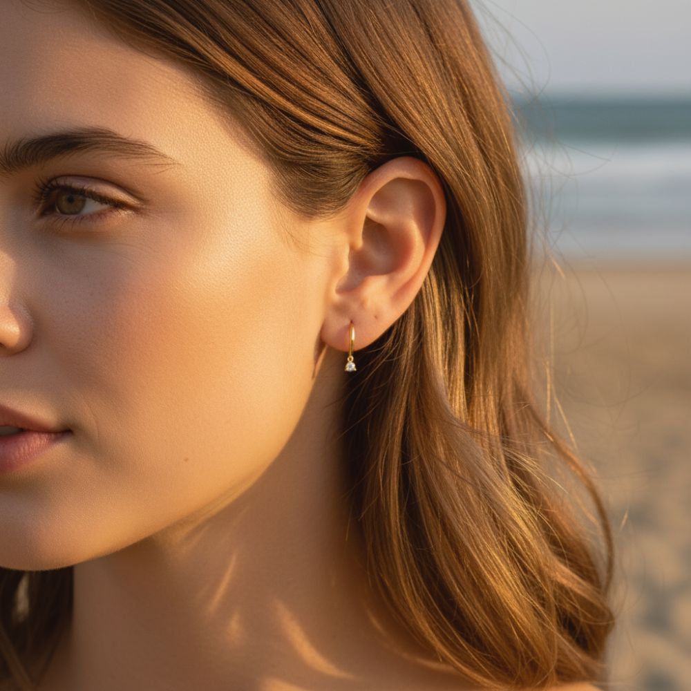Gold huggie earring with a dangling zircon stone worn on the ear in a close-up shot.