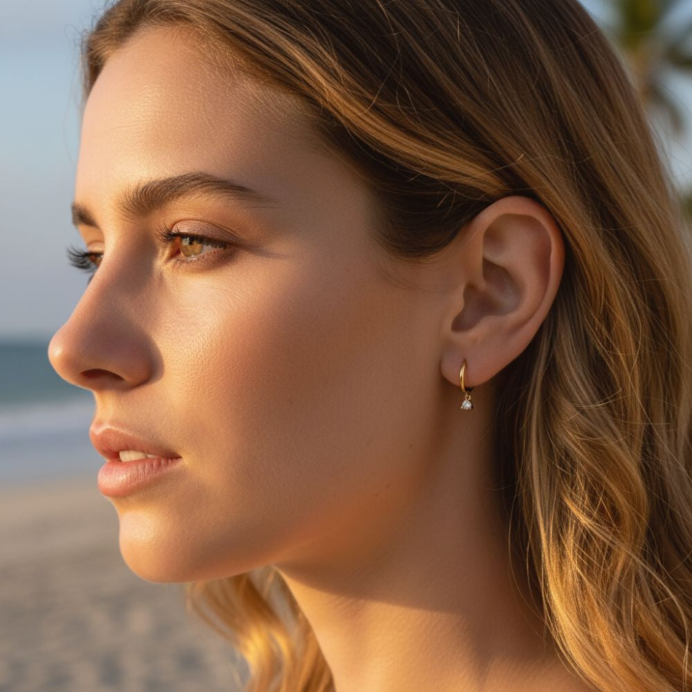 Gold huggie earrings with a dangling cubic zirconia stone worn on ear.