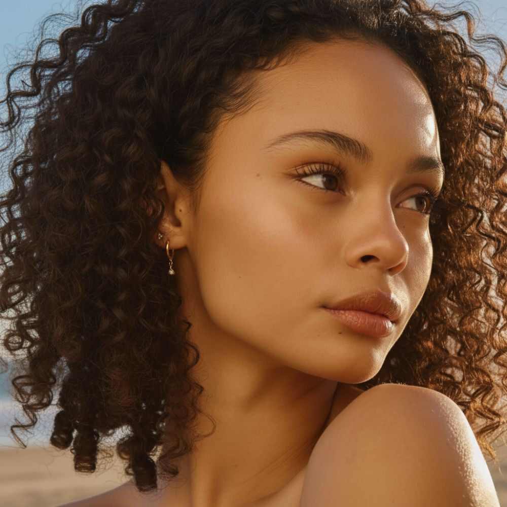 Model wearing gold huggie earrings with a small dangling stone on the beach.