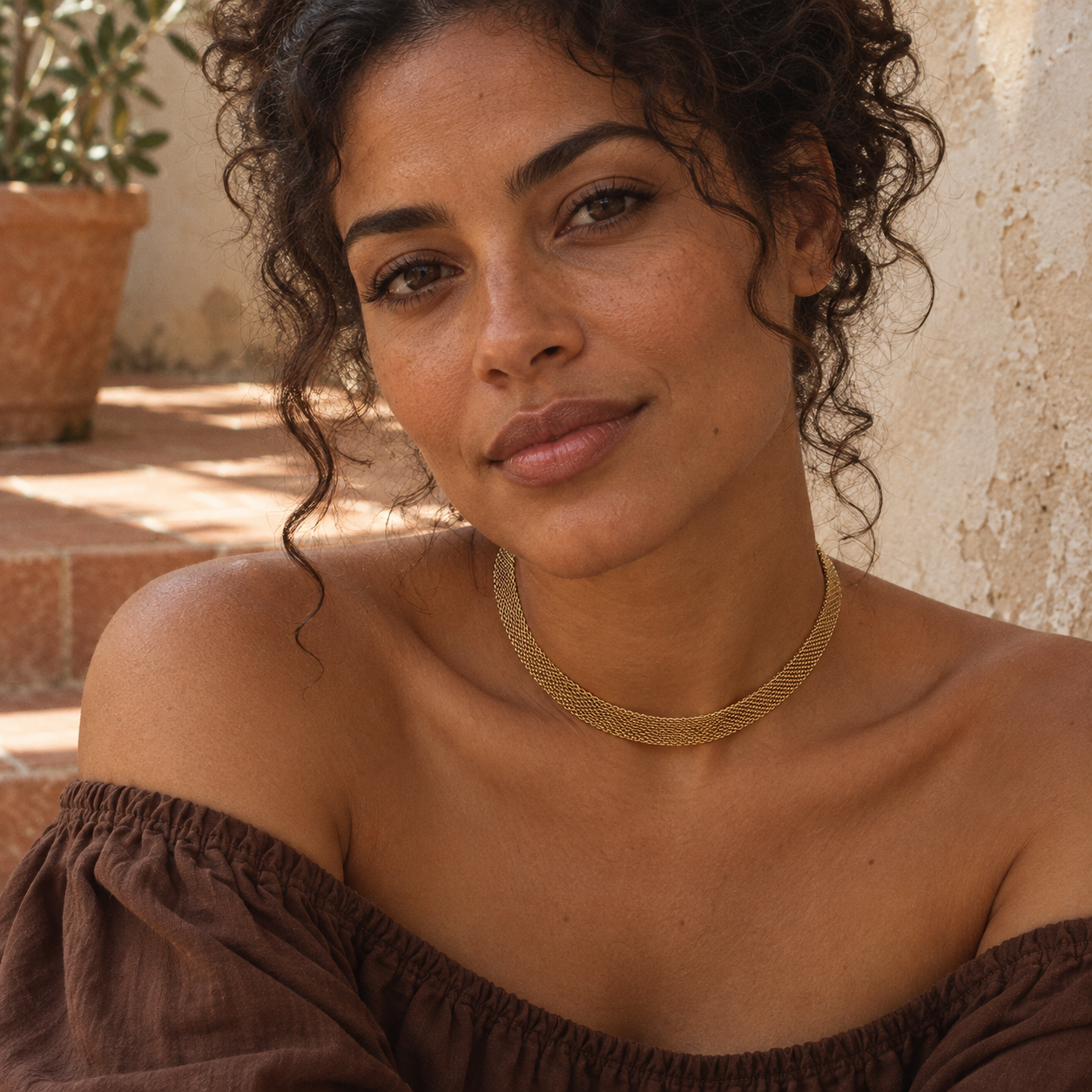 Woman wearing a gold mesh choker necklace with a brown off-the-shoulder top beside terracotta steps.