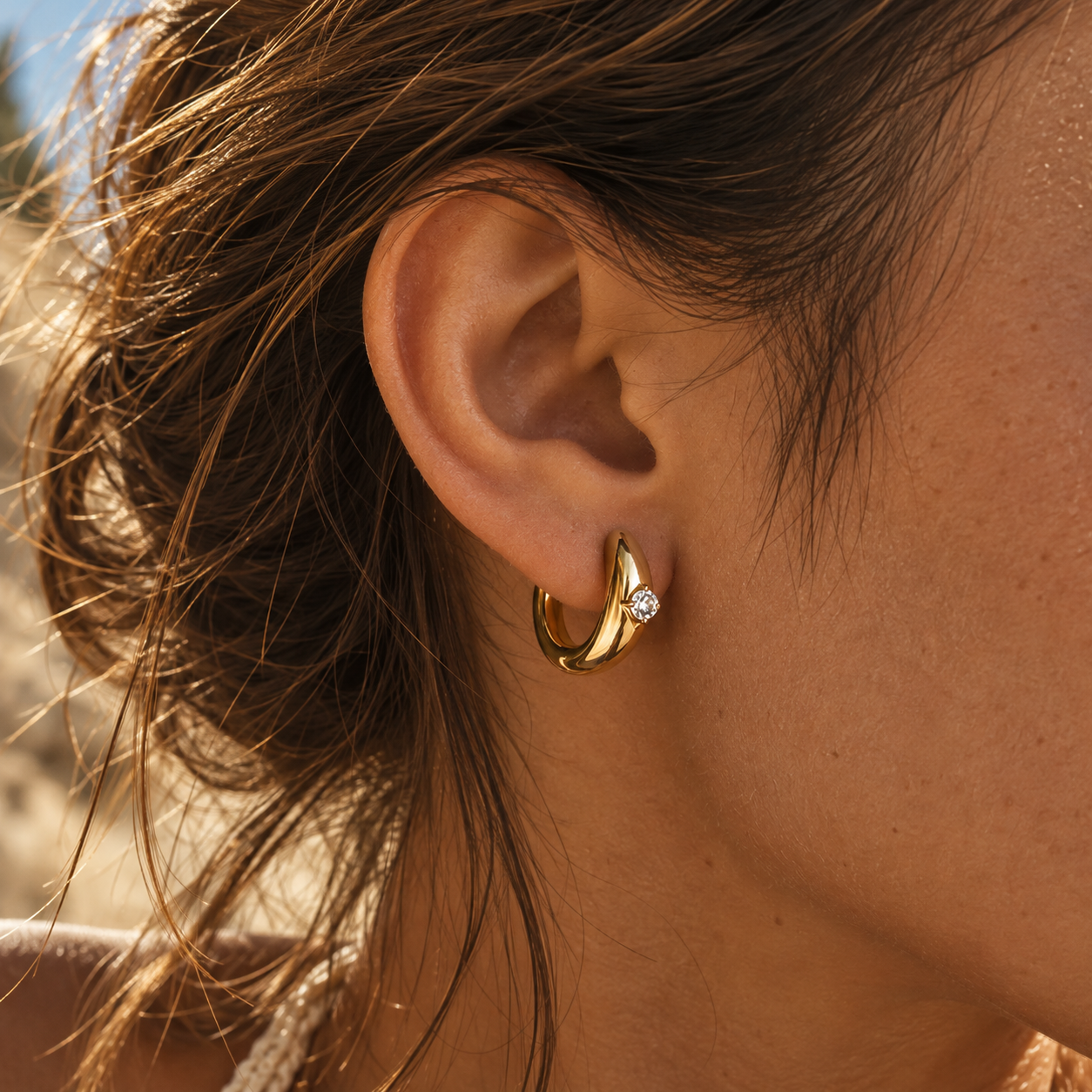 Gold zircon huggie earring worn on a woman’s ear, chunky gold-plated hoop with a sparkling stone, sunlit beach look