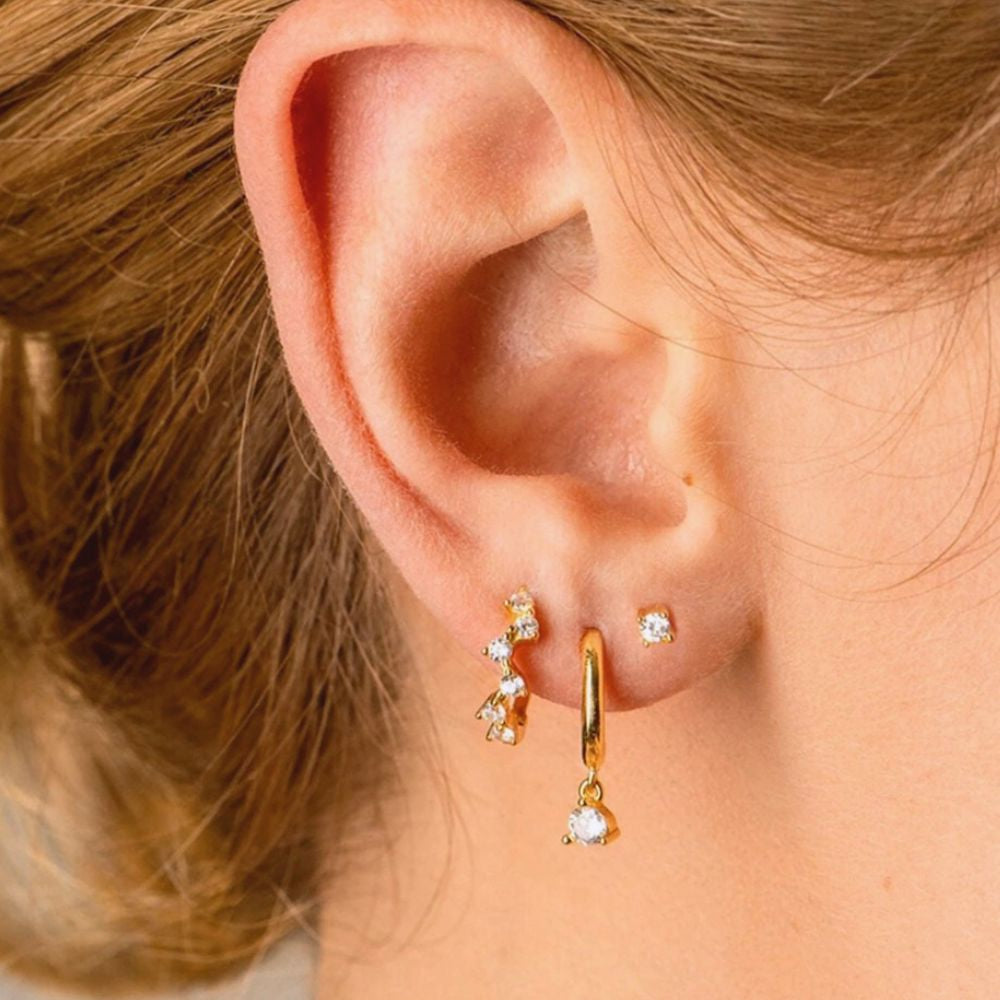 Gold huggie earrings with dangling zirconia stone displayed in a curated ear stack composition.