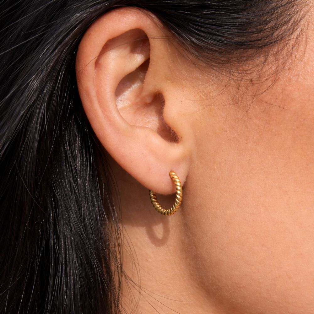 Close-up of gold twisted hoop earrings worn on ear, minimalist rope texture huggie hoops.