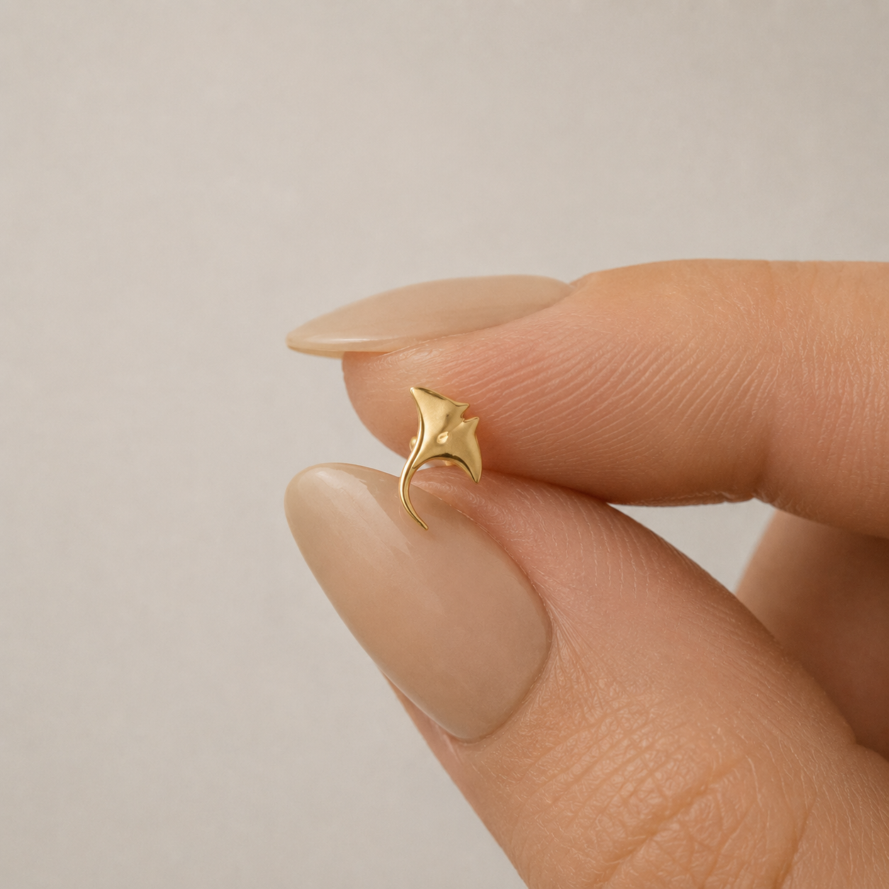 Tiny gold manta ray cartilage stud held between fingers with nude manicure