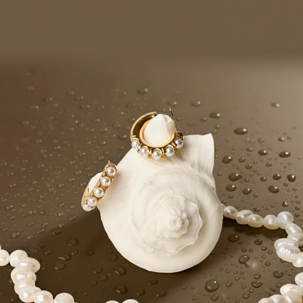Gold pearl huggie earrings displayed on a white seashell with pearls, beach-inspired waterproof jewelry