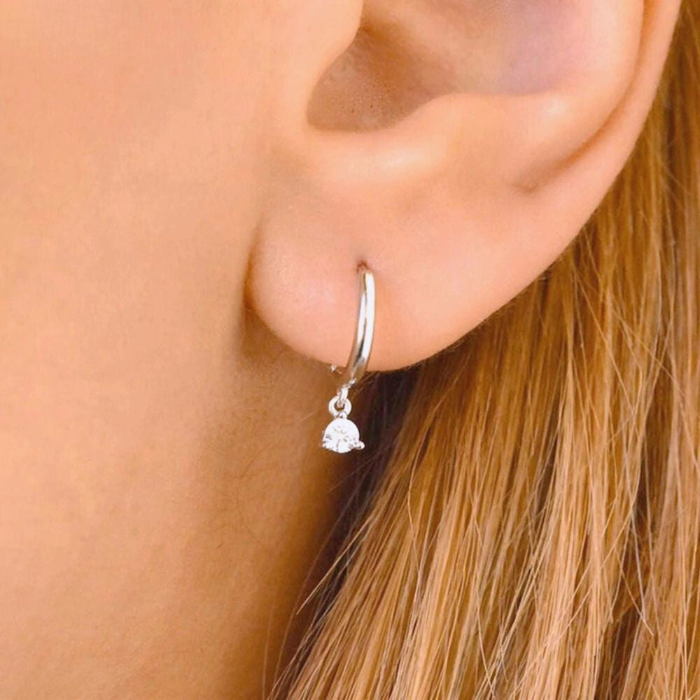 Close-up of a minimalist silver huggie earring with a dangling cubic zirconia stone.