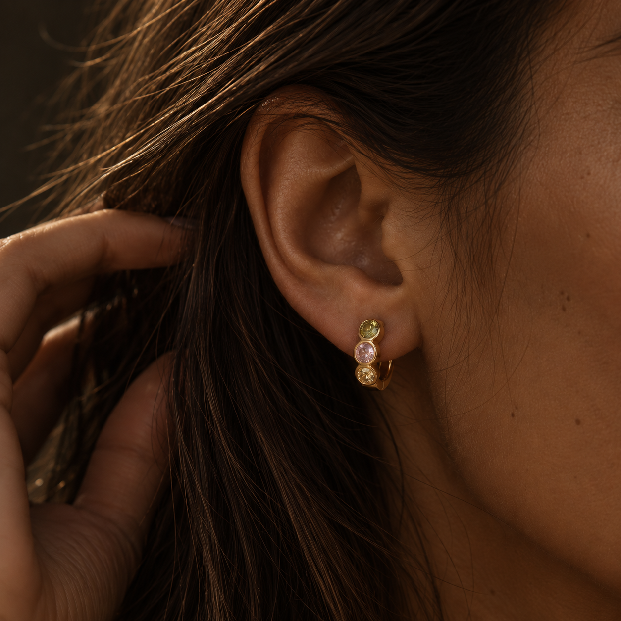 Colorful three stone gold huggie earring worn on ear in warm light