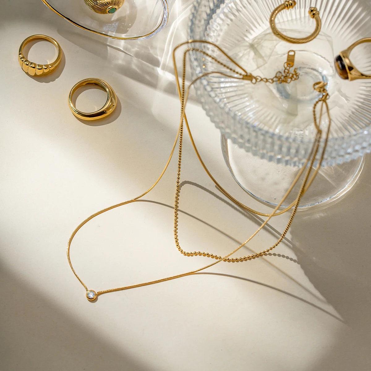 Zahia necklace styled with rings on a glass bowl under sunlight.